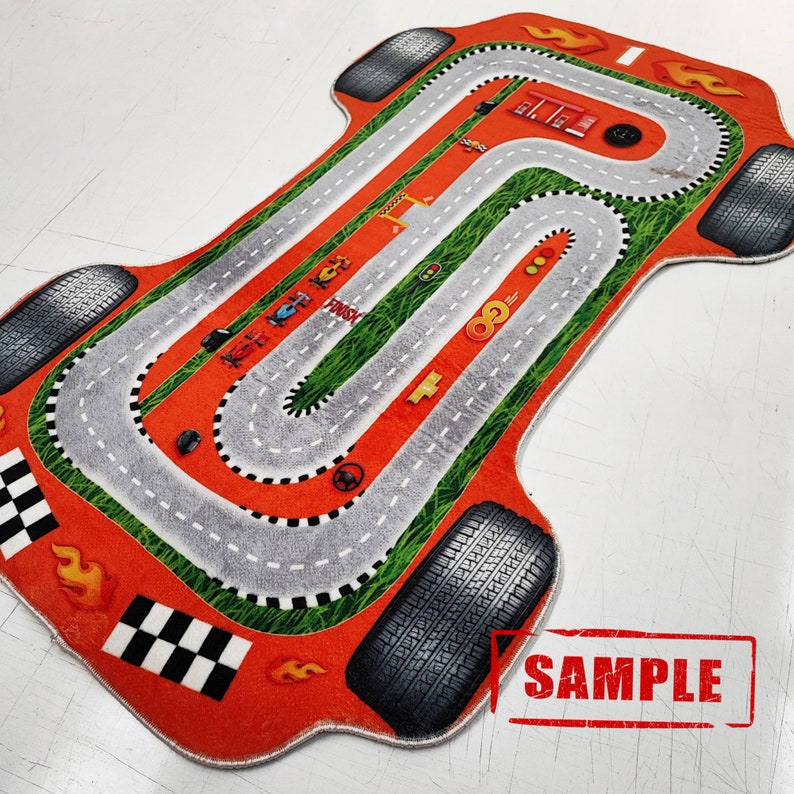 Puede incluir: Alfombra de pista de carreras de coches naranja y verde con una pista gris y una l&iacute;nea de meta a cuadros blanco y negro. La alfombra tiene forma de coche de carreras y tiene neum&aacute;ticos alrededor de los bordes.