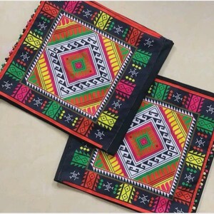 May include: Two square cushion covers featuring a black base with a vibrant geometric pattern in red, yellow, green, and white. The design includes concentric diamond shapes and intricate border motifs, finished with small colorful beads along the edges.