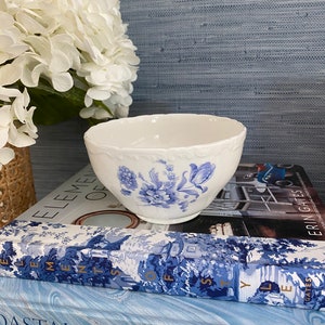 May include: A white porcelain bowl with blue floral patterns and a decorative rim. The bowl is placed on a stack of books with blue and white covers, and a bouquet of white flowers is in the background.