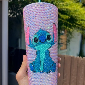 May include: A bedazzled tumbler featuring a blue cartoon character with large ears. The tumbler is covered in iridescent rhinestones, creating a shimmering effect. The character is blue with pink accents. A green straw is visible.