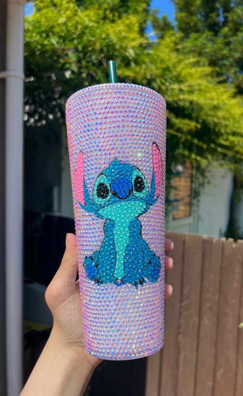 May include: A bedazzled tumbler featuring a blue cartoon character with large ears. The tumbler is covered in iridescent rhinestones, creating a shimmering effect. The character is blue with pink accents. A green straw is visible.