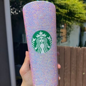 May include: A bedazzled Starbucks tumbler with a green straw. The cup is covered in iridescent rhinestones. The Starbucks logo is centered on the cup. The cup is held in front of a blurred background of green foliage.
