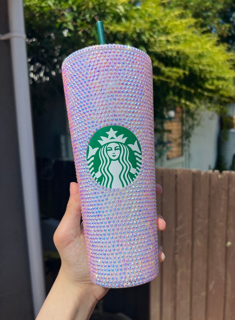 May include: A bedazzled Starbucks tumbler with a green straw. The cup is covered in iridescent rhinestones. The Starbucks logo is centered on the cup. The cup is held in front of a blurred background of green foliage.