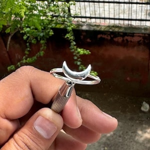 Può includere: Un anello d'argento con un design a mezzaluna. L'anello è tenuto in mano su uno sfondo sfocato di vegetazione e un muro marrone. La fascia dell'anello è sottile e la luna è liscia.
