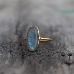 May include: A gold ring with an oval, blue-grey gemstone. The ring has a simple design with a thin gold band and a bezel setting. The gemstone has an iridescent quality. The ring is a piece of jewellery.