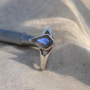 May include: A silver ring with a geometric design. The ring features a central, diamond-shaped stone with blue and purple iridescent hues. The ring is set against a neutral fabric background.