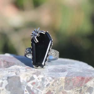 Puede incluir: Un anillo de plata con una piedra negra en forma de ataúd y una araña de plata en la parte superior.