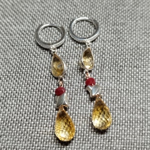 May include: Silver hoop earrings with a drop design. Each earring features a faceted, yellow citrine gemstone, a small red bead, and a silver geometric element. The earrings are designed to dangle and catch the light.