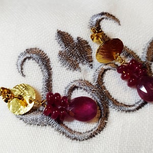 Op de afbeelding: Een paar gouden oorbellen met een ronde, getextureerde gouden schijf en een hangende cluster van dieprode kralen en een gefacetteerde, traanvormige rode edelsteen.