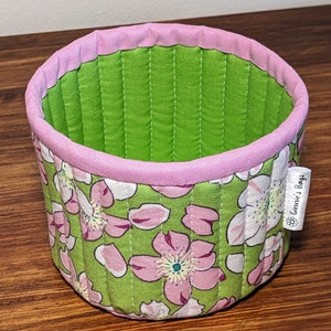May include: A green and pink fabric storage basket with a floral print and a label that reads "Ginnie's Bags".