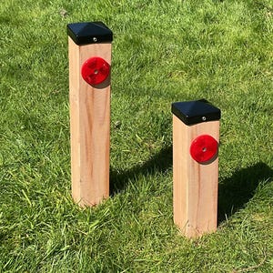 May include: Two wooden posts with black caps and red reflectors set in green grass. The posts are rectangular and made of light-coloured wood. The reflectors are circular and appear translucent.