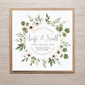 May include: A square wedding card featuring a watercolour floral wreath of white flowers and green leaves surrounding the text: CONGRATULATIONS, Aoife & Niall, Friday, 19th April, 2024, St. Brendan's Church, Birr, Co. Offaly, ON YOUR WEDDING DAY.