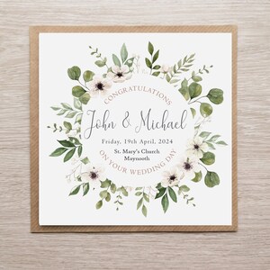 May include: A square wedding greeting card featuring a watercolour floral wreath of green leaves and white flowers. The text reads: CONGRATULATIONS John & Michael Friday, 19th April, 2024 St. Mary's Church Maynooth ON YOUR WEDDING DAY. The card has a brown border.
