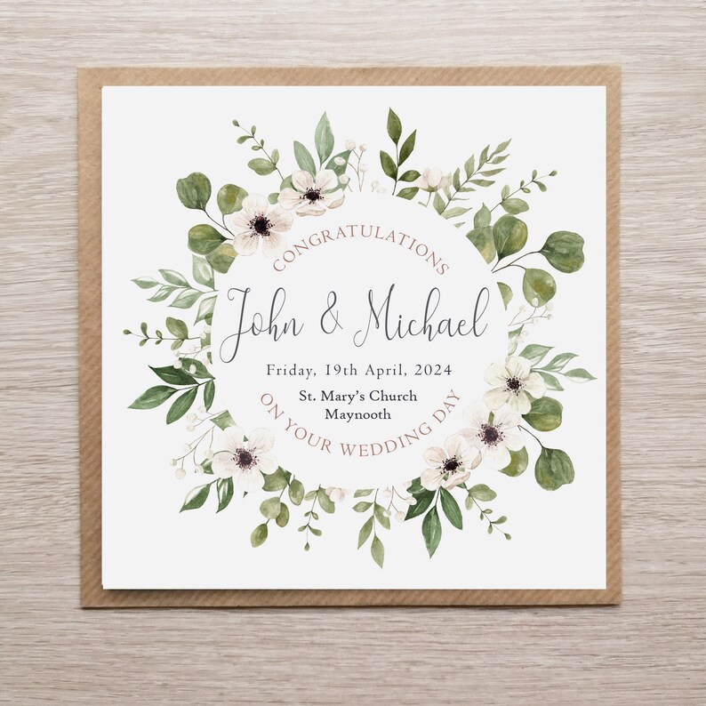 May include: A square wedding greeting card featuring a watercolour floral wreath of green leaves and white flowers. The text reads: CONGRATULATIONS John & Michael Friday, 19th April, 2024 St. Mary's Church Maynooth ON YOUR WEDDING DAY. The card has a brown border.