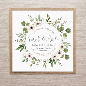 May include: A square wedding card featuring a watercolour wreath of green eucalyptus leaves and white flowers surrounding the text: CONGRATULATIONS, Sarah & Aoife, Friday, 19th April, 2024, St. Mary's Church, Maynooth, ON YOUR WEDDING DAY.