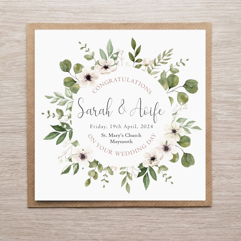 May include: A square wedding card featuring a watercolour wreath of green eucalyptus leaves and white flowers surrounding the text: CONGRATULATIONS, Sarah & Aoife, Friday, 19th April, 2024, St. Mary's Church, Maynooth, ON YOUR WEDDING DAY.