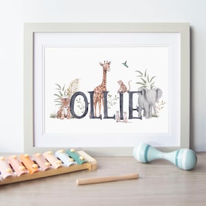Può includere: Una stampa artistica incorniciata che mostra il nome OLLIE in grandi lettere scure, integrata con illustrazioni ad acquerello di una tigre, una giraffa, una scimmia, suricati, un elefante e un colibrì, circondati da fogliame tropicale verde.