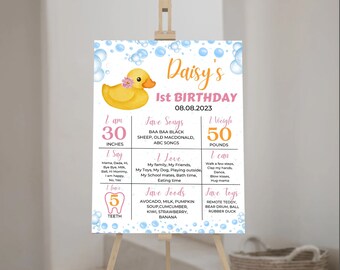 May include: A pink and blue first birthday milestone poster with a yellow rubber duck illustration. The poster lists the child's age, height, weight, favorite songs, foods, and toys. The text on the poster reads "Daisy's 1st Birthday 08.08.2023".