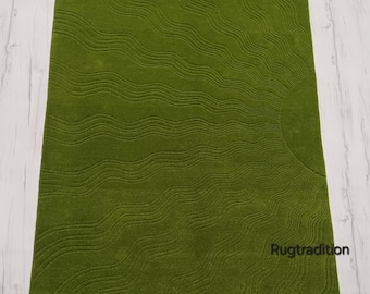 May include: A rectangular, olive green rug with a wave pattern design. The rug has a soft, textured surface and a sun-like design in the corner. The text "Rugtradition" is visible in the bottom right corner.