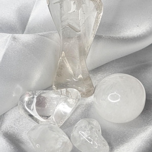 May include: A collection of clear quartz crystal figurines and stones. The assortment includes an angel figurine, a sphere, and several heart-shaped and irregular stones, all resting on a white satin surface.