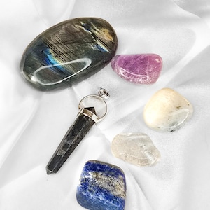 May include: An assortment of polished gemstones and a pendant on a white satin surface. The stones include a large, oval labradorite, a purple stone, a cream-colored stone, a clear stone, and a blue stone. The pendant is black with a silver-colored ring.