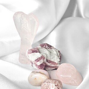 May include: A collection of crystals including a carved light pink rose quartz angel figurine, a cluster of raw pink tourmaline with white quartz, and three smooth, polished tumbled stones in shades of pale pink and translucent white.