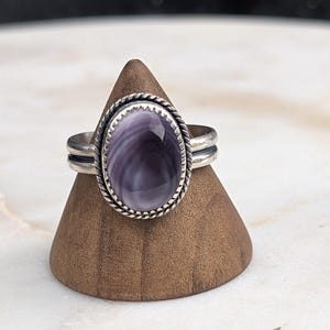 Könnte beinhalten: Ein Silberring mit einem großen, ovalen, lavendelfarbenen Stein. Der Stein ist in einer dekorativen Lünette mit einem gedrehten Seilmuster gefasst. Der Ring hat ein schlichtes Band mit zwei parallelen Linien. Der Ring wird auf einem Holzkegel präsentiert.