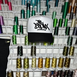 Puede incluir: Una gorra de béisbol blanca y negra con el texto "The Bronx" en letras negras estilo grafiti. La gorra se muestra frente a un estante lleno de carretes de hilo colorido, mostrando un tema de artesanía o bordado.
