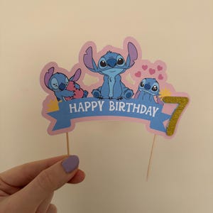 Puede incluir: Un adorno para pastel rosa y azul con tres personajes de Stitch de la pel&iacute;cula de Disney Lilo & Stitch. El adorno tiene una pancarta que dice "Feliz cumplea&ntilde;os" y un n&uacute;mero 7 de purpurina dorada.