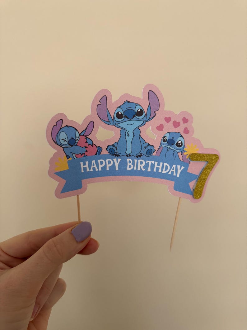 Puede incluir: Un adorno para pastel rosa y azul con tres personajes de Stitch de la pel&iacute;cula de Disney Lilo & Stitch. El adorno tiene una pancarta que dice "Feliz cumplea&ntilde;os" y un n&uacute;mero 7 de purpurina dorada.