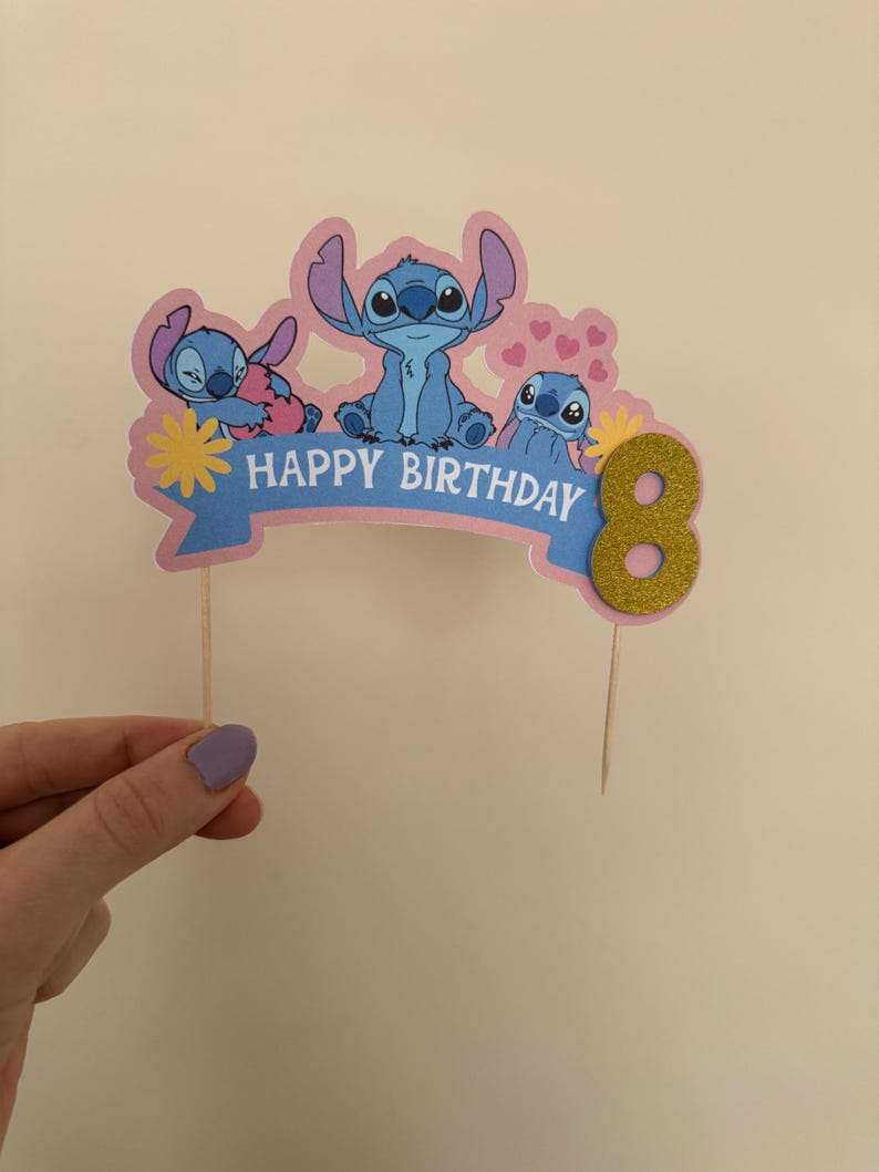 Puede incluir: Un adorno para pastel rosa y azul con tres personajes de Stitch de la pel&iacute;cula de Disney Lilo & Stitch. El adorno dice "Feliz cumplea&ntilde;os" y tiene un n&uacute;mero 8 de purpurina dorada.