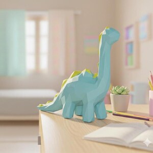 Puede incluir: Figura de dinosaurio geométrico azul claro con detalles amarillos. La figura está hecha de múltiples superficies planas, dándole una apariencia poligonal. Se encuentra sobre una superficie de madera, con una planta suculenta y lápices cerca.