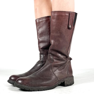 May include: A pair of knee-high, dark brown leather boots. The boots have a rounded toe, a low heel, and a side zipper. The leather appears worn, giving the boots a vintage look. The boots are standing on a white surface.