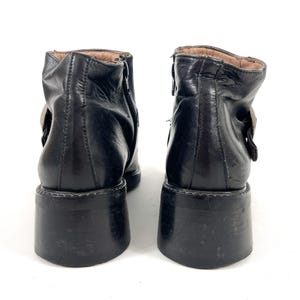 May include: A pair of black leather ankle boots with chunky platform soles. The boots have a side zipper and a buckle detail. The interior lining is a light brown color. The boots are set against a white background.