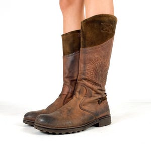 May include: Brown leather knee-high boots with a distressed finish. The boots feature a suede upper and a buckle detail. The boots have a round toe and a rubber sole. The boots are approximately 14 inches tall.