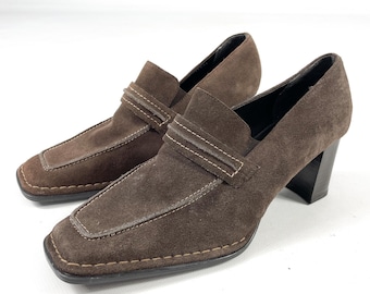 May include: A pair of brown suede heeled loafers. The shoes feature a square toe, a strap across the top, and a stacked heel. Visible stitching details the edges. The background is white.