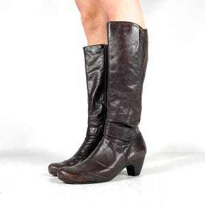 May include: A pair of dark brown leather knee-high boots. The boots have a rounded toe, a small heel, and a strap with a button detail around the ankle. The boots have a zipper on the side.
