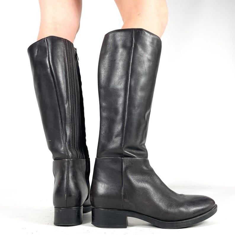 May include: A pair of black leather knee-high boots. The boots have a rounded toe and a low heel. One boot has a zipper on the side. The boots are made of a smooth, shiny leather material.