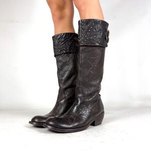 May include: Dark brown mid-calf boots featuring a distressed, wrinkled leather texture. The boots have a wide, folded-over cuff at the top adorned with small metallic studs and two side buckles, finished with a rounded toe and a low block heel.