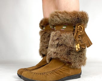 May include: Brown suede boots with faux fur trim. The boots have a moccasin-style toe and a decorative band with geometric patterns. Tassels and wooden beads add detail to the side of the boots. The boots appear to be ankle-high.
