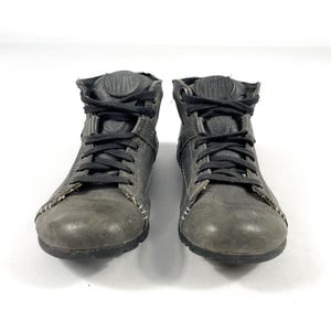 May include: Pair of gray leather ankle boots with black laces. The boots have a worn appearance, with visible stitching and a textured finish. The design includes a padded collar and a rounded toe. The boots are set against a white background.