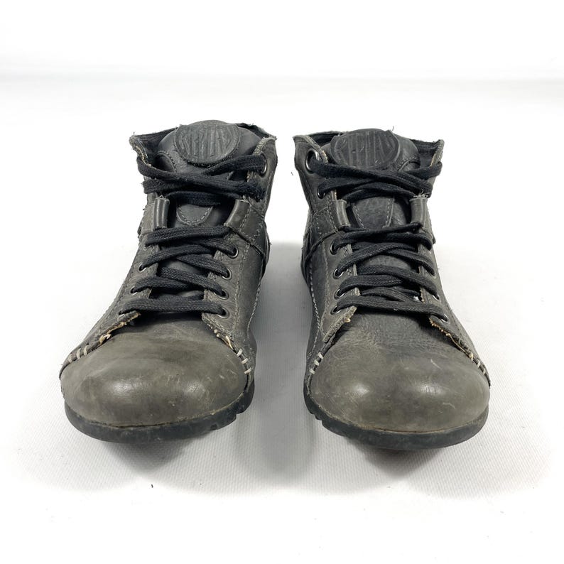 May include: Pair of gray leather ankle boots with black laces. The boots have a worn appearance, with visible stitching and a textured finish. The design includes a padded collar and a rounded toe. The boots are set against a white background.
