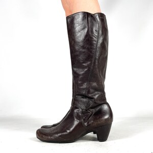 May include: A pair of brown leather knee-high boots. The boots have a rounded toe, a small heel, and a strap with a button detail around the ankle. The leather has a slightly distressed appearance.