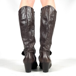 May include: A pair of brown leather knee-high boots. The boots have a slight heel, a zipper on the side, and a buckle detail. The leather appears worn, giving the boots a vintage look. The boots are shown from the back.