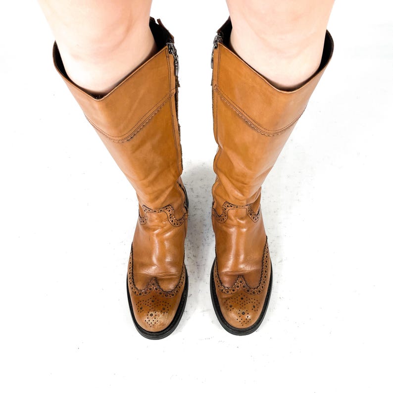 May include: A pair of knee-high tan leather boots featuring brogue detailing on the rounded toe and vamp. The boots have a vertical zip closure along the inner side and a black sole, providing a structured look with visible stitching accents.