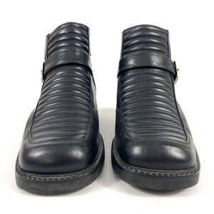 May include: Pair of black leather ankle boots with a square toe and a ridged design. Each boot features a strap with a silver buckle. The boots have a thick, textured sole.