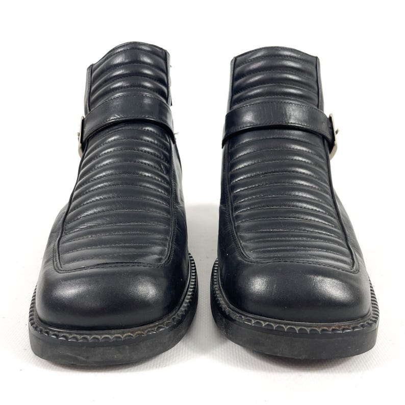 May include: Pair of black leather ankle boots with a square toe and a ridged design. Each boot features a strap with a silver buckle. The boots have a thick, textured sole.