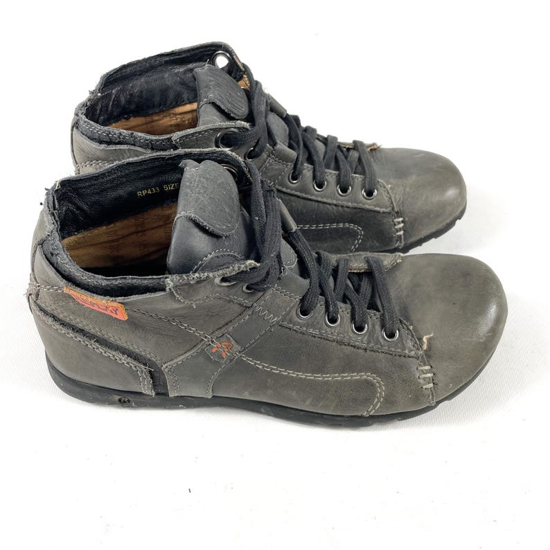 May include: A pair of gray leather ankle boots with black laces. The boots have a distressed look with visible stitching and a small orange accent. The interior of the boots is visible, and the soles are dark.