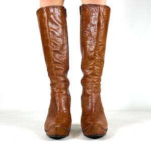 May include: A pair of knee-high brown leather boots. The boots have a pointed toe, a zipper on the side, and a stitched design around the top and toe. The leather has a textured appearance.