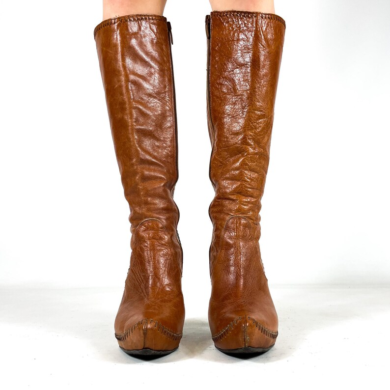 May include: A pair of knee-high brown leather boots. The boots have a pointed toe, a zipper on the side, and a stitched design around the top and toe. The leather has a textured appearance.